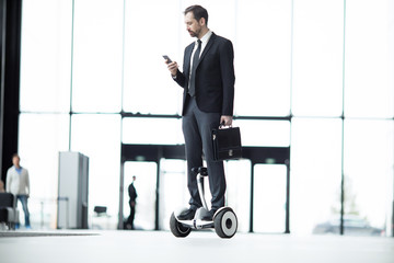 Young busy employee with briefcase texting in smartphone while moving along modern center on hoverboard