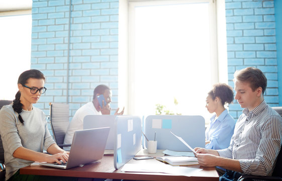 Four Young Managers Of Open Space Office Sitting By Individual Workplaces And Carrying Out Organization Work