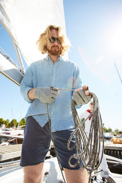 Young man in blue shirt, shorts, sunglasses and gloves preparing rope before riding on yacht