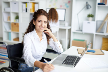 Happy young businesswoman in wheelchair looking at you by her workplace in office