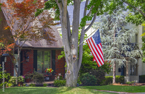 Brick house set in traditional neighborhood with large trees a bird feeder and colorful flowers and an American flag