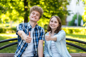 Fototapeta premium Young couple girl and boy. Summer in park on a bench. Smiles happily. Hard hands showing thumbs up, like. Emotionally gestures. The concept of success and success.