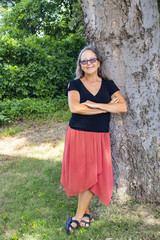 Woman with long grey hair and glasses and organge skirt and black blouse leans against large tree with arms crossed and smile on her face in summer
