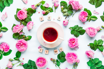 Top view creative layout with Tea time lettering with wooden blocks, cup of hot tea and fresh pink tea rose flowers, buds, petals, leaves on white background isolated. Flat lay. Copy space.