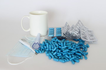 Treatment, medicine - a lot of blue capsules on the background of a white mug, digital thermometer, medical mask, blister with tablets
