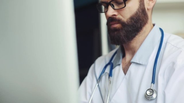 Brown-haired Caucasian physician in spectacles sitting at table and looking at monitor. Content man with beard typing on keyboard of computer at clinic. Indoors.