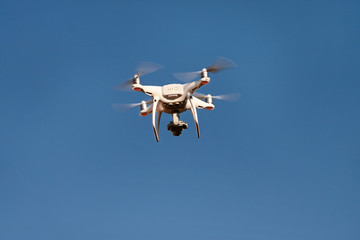 Drone quad copter fly on blue sky in background. Modern drone is flying in air, to take photos and record footage from above. Drone with four propellers, camera and red warning lights. New technology.