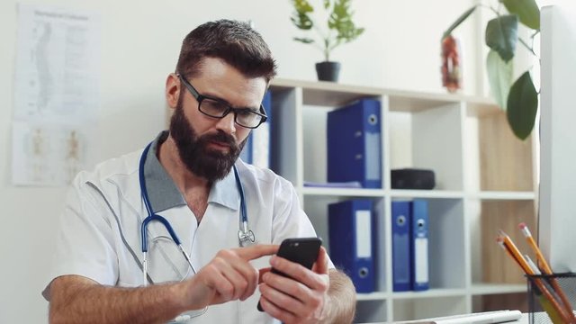 Good-looking man wearing white lab coat at doctor’s office. Caucasian doctor using smartphone, browsing on background of shelves with blue folders. Indoors. Clinic.
