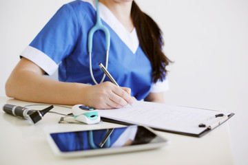 Female doctor with otoscope and pulse oximeter in blue uniform making notes in application form. Medicine and health care concept.