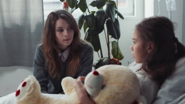 Mom and daughter speking at hospital room. Young Ukrainian unhappy mother sitting near hospital bad with girl. Conversation.