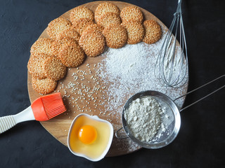 Top view of product set for cooking cookies, kitchenware and several round cookies with sesame on a black wooden table.