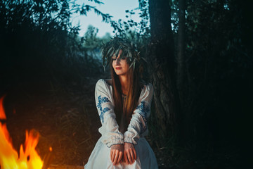 Woman in ethnic clothes with chaplet on a head sitting near bonfire. Concept of Pagan holiday.