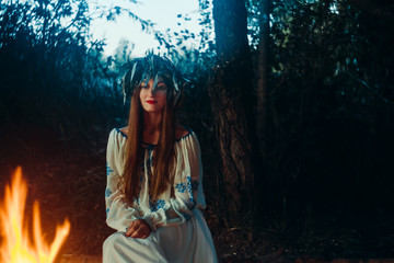 Woman in ethnic clothes with chaplet on a head sitting near bonfire. Concept of Pagan holiday.