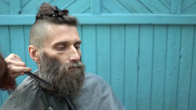 Barber woman carefully trimming beard of client with clipper and comb at outdoor barbershop