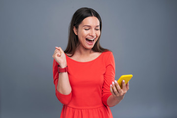 Fantastic news. Charming young woman being happy about the message, having read the message on her smartphone, while standing isolated on a blue-grey background