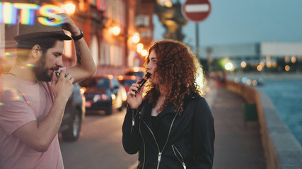 Loving couple of hipsters on date. Guy in hat and t-shirt smokes electronic cigarette. Girl with curly red hair is holding ecigarette.