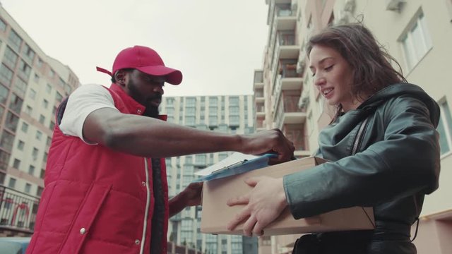 On street courier African American gives a parce to a woman after signing on delivery device cardboard technology box order post postal service device sunny beard covered load logistic mailman