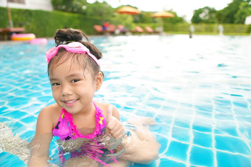 Asian children or kid girl wear pink bikini and goggles with thumb up for like and good or great on swimming pool or water park and smile with happy for refreshing and relax exercise on summer holiday