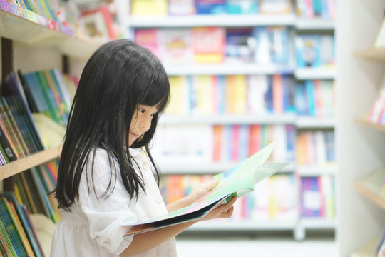 Asian children cute or kid girl smile reading and choose tale or story book on bookshelf in bookstore or library room at school or nursery for learning and studying with education