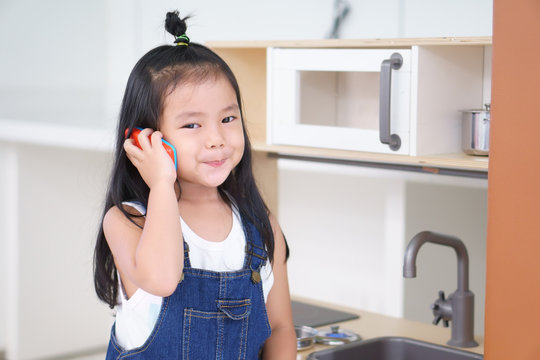 Asian Children Cute Or Kid Girl Smile And Playing Smartphone Toy Or Talking On Phone In Kitchen Room And Kitchenware Toy For Communication And Cooking Food Learning And Housewife With Wear Dungarees