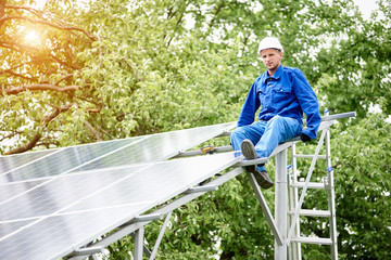 Young smiling electrician points screwdriver at camera sitting on almost finished stand-alone solar photo voltaic panel system on bright sunny green tree background. Alternative energy concept.
