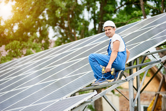 Young smiling electrician worker sitting on almost finished stand-alone solar photo voltaic panel system on bright sunny green tree background. Alternative energy concept.