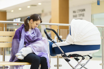 Young european woman with violet stole is breastfeeding her little child close to white baby carriage at public place shopping mall
