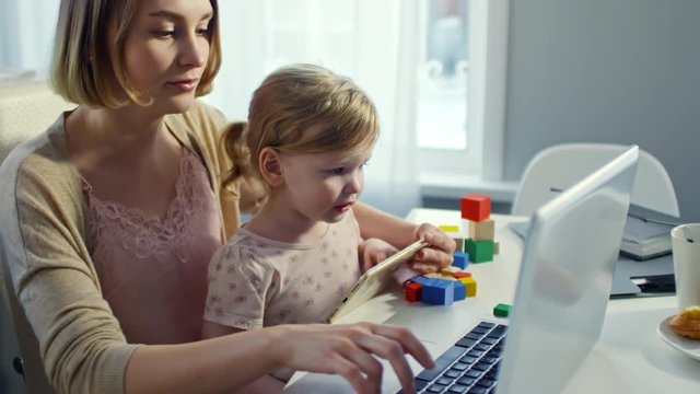 PAN of busy young mother holding cute toddler girl on her laps and working on laptop at home