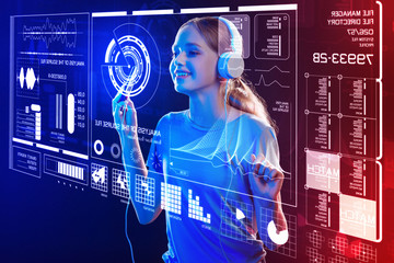 Leisure time. Cheerful emotional schoolgirl standing in front of a transparent screen and feeling happy while listening to music in her big headphones