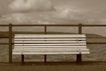 Fototapeta na wymiar Pusta drewanian ławka na molo, w tle morze (jezioro), linia brzegowa, pochmurne niebo, sepia