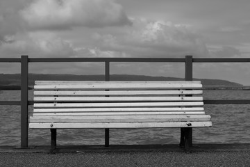 Pusta drewniana biała ławka stoi pusta na brzegu jeziora (nabrzeżu morza), za nią niska metalowa barierka, w tle woda, horyzont, chmury na niebie, czarno-białe - obrazy, fototapety, plakaty