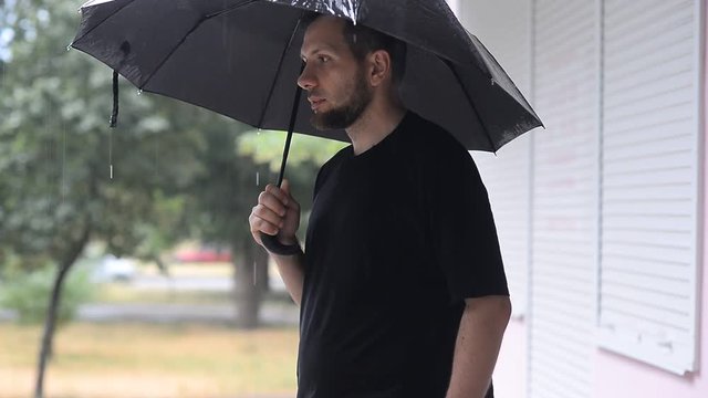 Bearded man in black shirt stands under the umbrella in the rain and sneezes