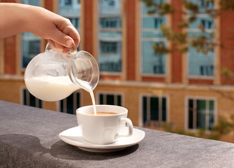 Tea with milk in the morning on the balcony. Milk is poured into a cup of tea. Tea with milk on the windowsill the balcony in the morning on the background of windows of the house.
