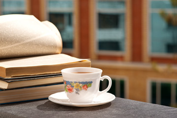 A cup of tea and books on the windowsill. A cup of tea on the balcony window sill in the sun. The wind turns over the pages of the book. Morning tea.