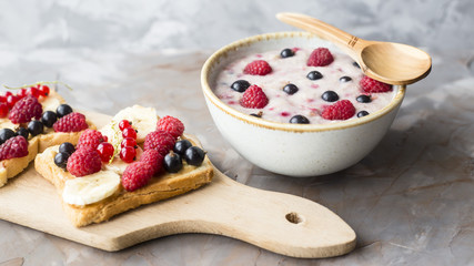 A bowl of oatmeal with berries next to a board with sandwiches with peanut butter and banana, sporodina, raspberries. Healthy summer breakfast on a gray table