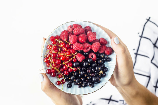 Berries on a plate in female hands on a white background. Black and red currants and raspberries. Next to a black and white checkered cloth. Top view, flat lay