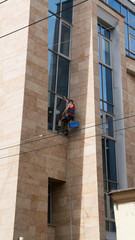 A worker is hanging on ropes and doing works for cleaning and washing of a buildings facade. Sunny warm day. Picture is made from a ground.