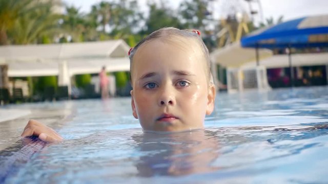 funny and sad child swims in the pool, the girl looks with big eyes at the camera