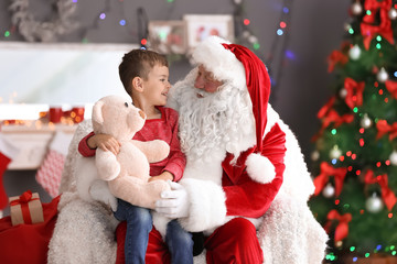 Fototapeta na wymiar Little boy with teddy bear sitting on authentic Santa Claus' lap indoors