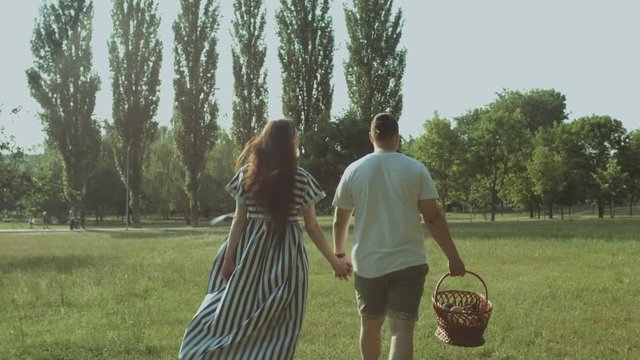 Back view of cheerful young family holding hands, carrying picnic basket and going on summer picnic in rays of setting sun. Cheerful couple enjoying leisure and walking in public park.