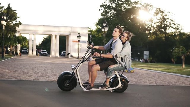 Confident man in blue shirt driving scooter while his girlfriend embracing him. Amazing young woman riding on moped with friend by the parkside