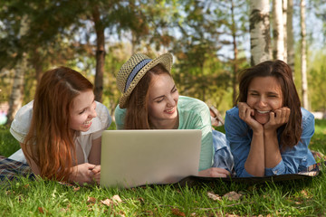 group of young teen girls are lying on a summer green lawn with a laptop and laughing. The concept of outdoor recreation, warm weather and pleasant holidays