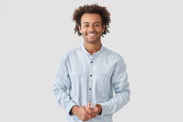 Horizontal shot of happy mixed race male with shining smile, feels glad as recieves bonus and praise for good work, has specific appearance, Afro hairstyle, dressed in fashionable white shirt - obrazy, fototapety, plakaty