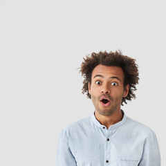 Puzzled handsome mixed race young male looks with surprised expression, opens mouth in bewilderment, exclaims in shock, has scared look, crisp hairstyle, dark skin isolated over white blank wall