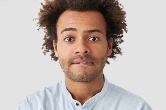 Close up portrait of attractive young hipster looks embarrassed, bites lips, looks seriously at camera, has bushy hairstyle. Handsome mixed race man raises eyebrows in bewilderment, isolated