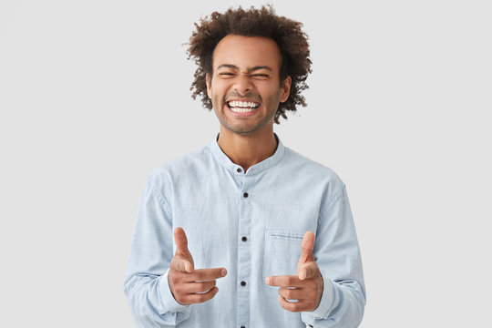 Positive mixed race male startup owner rejoices success, points at camera, has broad smile, being in high spirit, dressed in casual shirt, stands at studio. Confident student happy to graduate