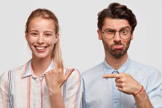 Positive young glad European female with blonde hair indicates with thumb at her boyfriend who has cranky facial expression, stand closely against white background, express different emotions