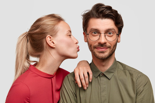 Affectionate girlfriend with light hair combed in pony tail, going to kiss her boyfriend, demonstrate truthful love, dressed in fashionable clothes, isolated over white background. People, relations