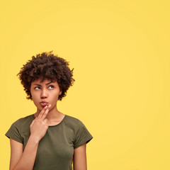 Pensive dark skinned female with Afro hairstyle, has serious expression, holds chin and being focused upwards, dressed in casual t shirt, poses against yellow background with blank copy space