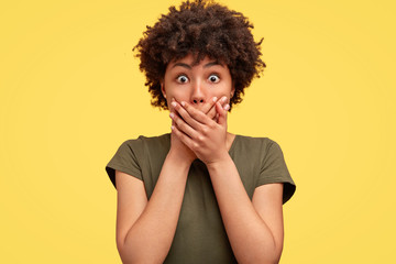 Emotive stupefied young African American female with dark skin, covers mouth as tries to be speechless, looks with popped eyes, stands against yellow background, shocked as notices car accident
